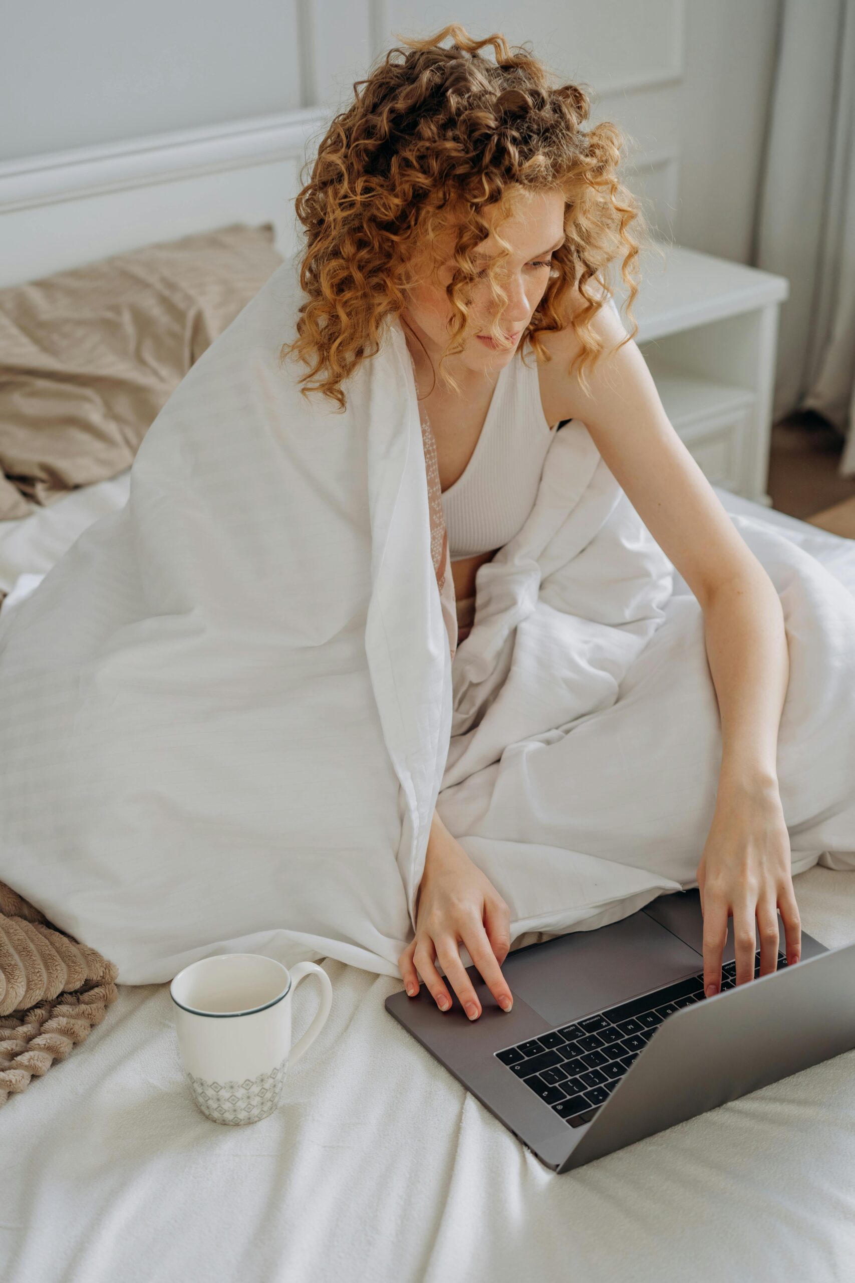 A woman with curly hair sitting on a bed using a laptop, wrapped in a blanket, with a cup nearby.