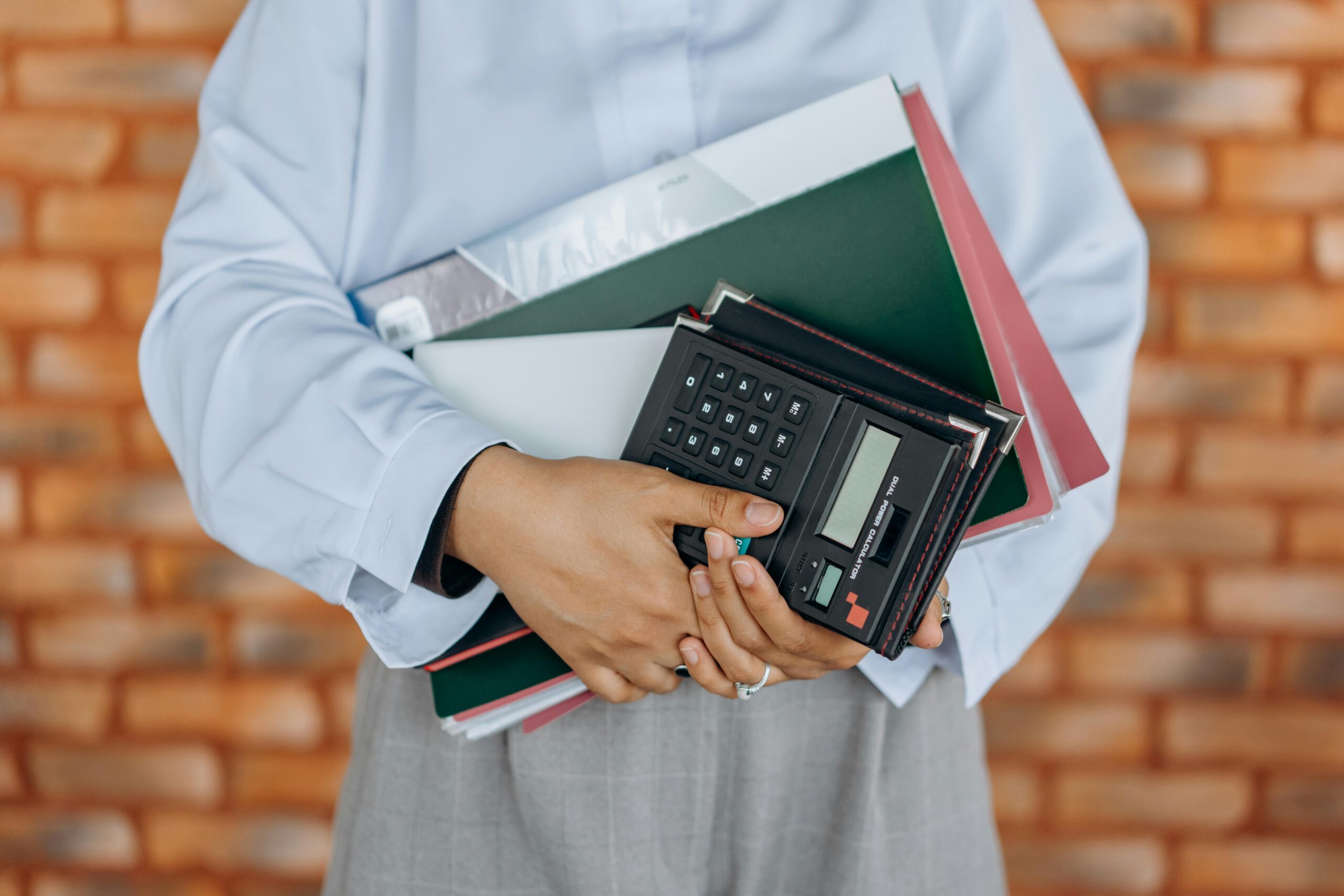 Close-up of a person holding a calculator and folders against a brick wall background.